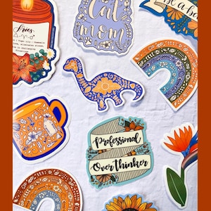 Pu&ograve; includere: Una collezione di adesivi colorati con vari design, tra cui una candela, una tazza, un dinosauro, un arcobaleno, un girasole e uno striscione che dice "Professional Overthinker". Gli adesivi sono tutti su uno sfondo bianco. Il testo "NEW DESIGNS CREATED" &egrave; in alto nell'immagine, e il testo "EVERY MONTH" &egrave; in basso.