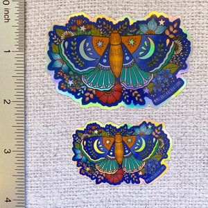 May include: Two iridescent holographic stickers featuring a blue, green, and orange moth with crescent moons and stars surrounded by flowers. The stickers are on a white fabric background with a ruler in the left corner. The text "@michaelakcreations" is on both stickers.
