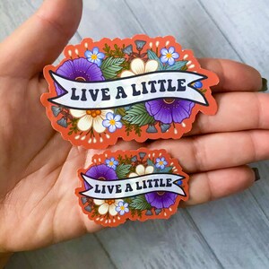 May include: Two floral stickers with the text "Live a Little" in a white banner. The stickers are on a peach background with purple, white, and blue flowers.