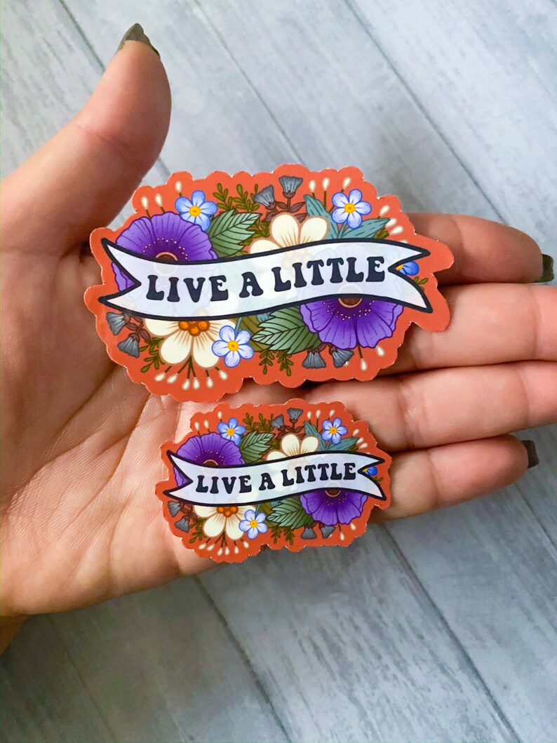 May include: Two floral stickers with the text "Live a Little" in a white banner. The stickers are on a peach background with purple, white, and blue flowers.
