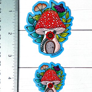 Puede incluir: Dos pegatinas de casa de seta con fondo azul. La pegatina m&aacute;s grande mide 6,5 cm de alto y la m&aacute;s peque&ntilde;a 4 cm de alto. Ambas pegatinas representan una seta roja y blanca con una puerta y una flor en la parte delantera. La seta est&aacute; rodeada de hojas verdes y flores.