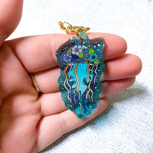 K&ouml;nnte beinhalten: Ein blauer und lilafarbener Quallen-Anh&auml;nger mit floralen Details und einer goldenen Kette. Der Anh&auml;nger ist aus Harz gefertigt und hat eine glitzernde Oberfl&auml;che.