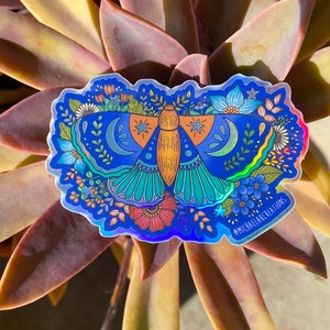 May include: A holographic sticker featuring a colorful moth with intricate details, surrounded by flowers and stars. The sticker has a blue background and the text "@michaelflakcreations" in the bottom right corner.