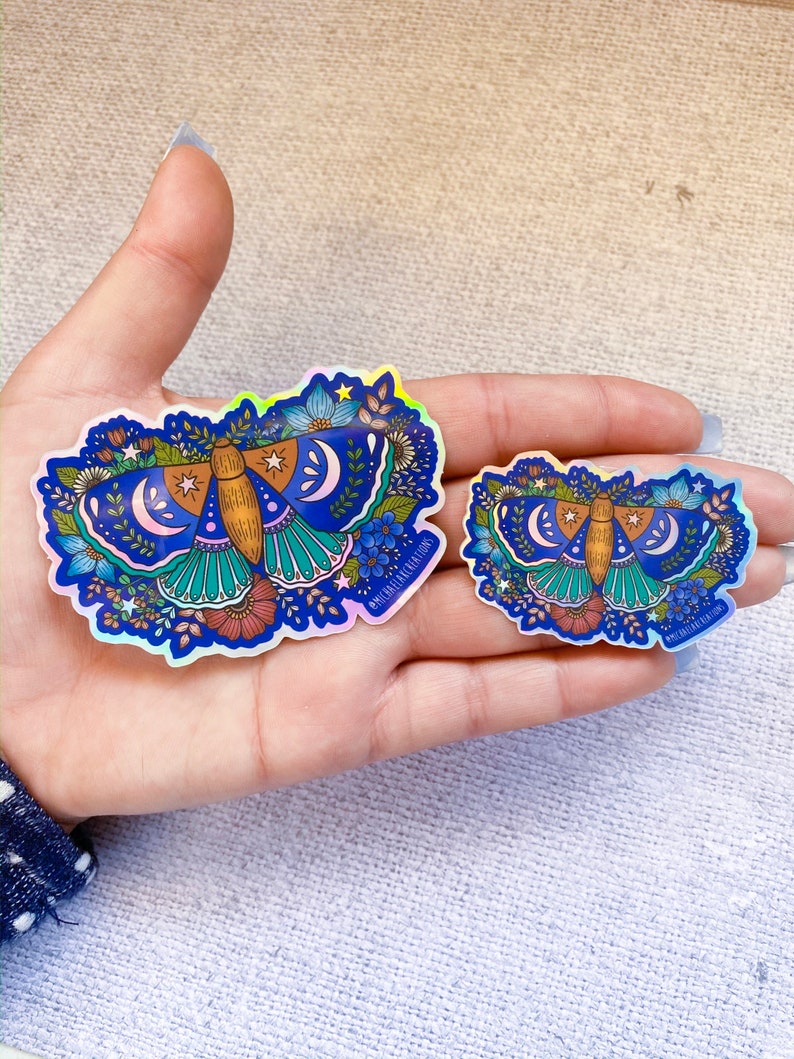 May include: Two iridescent holographic stickers of a blue and brown moth with floral details and crescent moons. The stickers are on a hand, with the larger sticker on the left and the smaller sticker on the right. The text "#michaelainkcreations" is on the bottom right corner of each sticker.