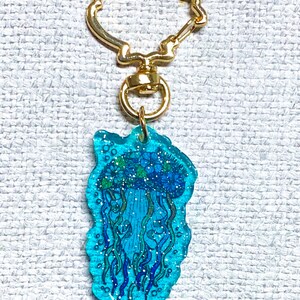 K&ouml;nnte beinhalten: Ein blauer und gr&uuml;ner Quallen-Schl&uuml;sselanh&auml;nger mit Glitzer, der in das Harz eingebettet ist. Der Schl&uuml;sselanh&auml;nger ist an einem goldfarbenen Metallring befestigt.