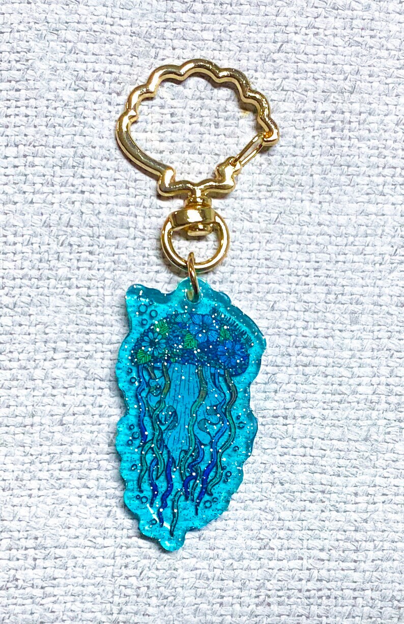 K&ouml;nnte beinhalten: Ein blauer und gr&uuml;ner Quallen-Schl&uuml;sselanh&auml;nger mit Glitzer, der in das Harz eingebettet ist. Der Schl&uuml;sselanh&auml;nger ist an einem goldfarbenen Metallring befestigt.