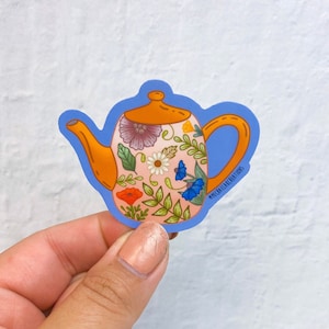 May include: A sticker of a floral teapot with a light pink background, orange spout, and a blue outline. The teapot is decorated with flowers, leaves, and a small blue bird. The sticker has the text "@michellaracraftings" in the bottom right corner.