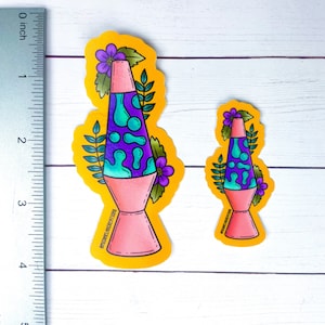 May include: Two colorful lava lamp stickers on a yellow background. The larger sticker is approximately 3 inches tall and features a pink lava lamp with purple and blue lava, green leaves, and purple flowers. The smaller sticker is approximately 2 inches tall and features a similar design. Both stickers have the text "Enchanted Ink Creations" at the bottom.