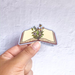 May include: A sticker of an open book with a bouquet of flowers growing from the pages. The sticker has a white background and the text "@michaelflaxcreations" is printed on the bottom.