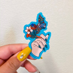May include: A blue sticker with a hand holding a bouquet of flowers. The flowers are in shades of red, orange, yellow, white, and blue. The hand has green nail polish. The sticker has the text "WILD BLACK CREATIONS" on the bottom.