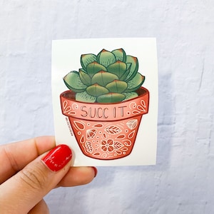 May include: A sticker featuring a green succulent plant in a pink pot with white floral designs. The pot has the text "SUCC IT." written on it.