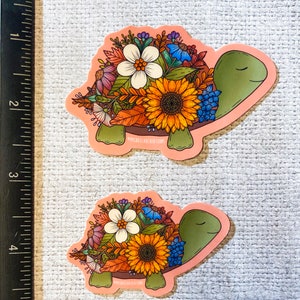 May include: Two pink floral turtle stickers. The turtles are outlined in black and have green shells. The flowers are a mix of yellow, orange, white, blue, and pink.