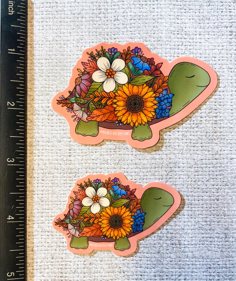 May include: Two pink floral turtle stickers. The turtles are outlined in black and have green shells. The flowers are a mix of yellow, orange, white, blue, and pink.