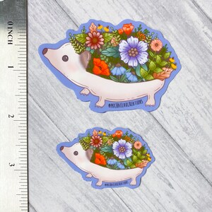 May include: Two stickers featuring a cartoon hedgehog with a floral arrangement on its back. The hedgehog is white with a light purple outline and a variety of colorful flowers. The text "MICHAELAXCREATIONS" is printed on the bottom of each sticker.