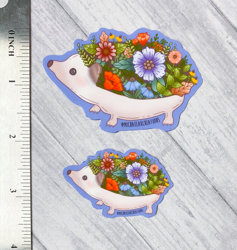 May include: Two stickers featuring a cartoon hedgehog with a floral arrangement on its back. The hedgehog is white with a light purple outline and a variety of colorful flowers. The text "MICHAELAXCREATIONS" is printed on the bottom of each sticker.
