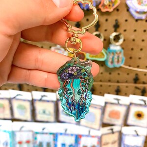 K&ouml;nnte beinhalten: Ein goldener Schl&uuml;sselanh&auml;nger mit einem blauen und violetten Quallen-Anh&auml;nger. Die Qualle ist von Blasen umgeben und hat ein Blumenmuster.