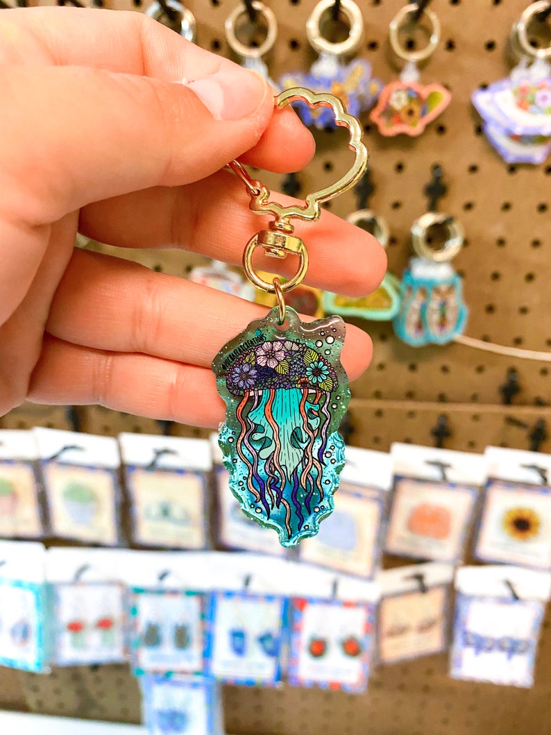 K&ouml;nnte beinhalten: Ein goldener Schl&uuml;sselanh&auml;nger mit einem blauen und violetten Quallen-Anh&auml;nger. Die Qualle ist von Blasen umgeben und hat ein Blumenmuster.