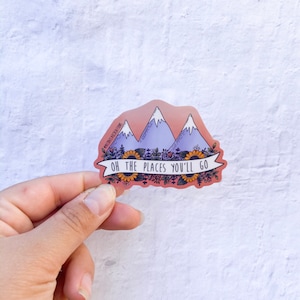 May include: A sticker with a pink background featuring three purple mountains with snow-capped peaks. The mountains are surrounded by yellow sunflowers and green leaves. The text "OH THE PLACES YOU'LL GO" is written in black on a white banner.