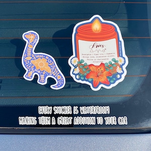 May include: Two waterproof car stickers. One sticker features a blue and orange dinosaur with floral patterns. The other sticker features a candle with a red jar and a blue rim, with a floral design and the text "Aries &Delta;.&gamma;.&sigma; Eucalyptus | Tiger lily | Chamomile Brings love, eliminates stress, Thrive in challenging situations".