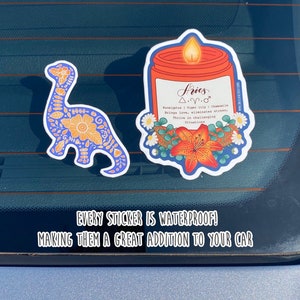 Pu&ograve; includere: Due adesivi impermeabili per auto. Un adesivo presenta un dinosauro blu e arancione con motivi floreali. L'altro adesivo presenta una candela con un barattolo rosso e un bordo blu, con un design floreale e il testo "Aries &Delta;.&gamma;.&sigma; Eucalyptus | Tiger lily | Chamomile Brings love, eliminates stress, Thrive in challenging situations".