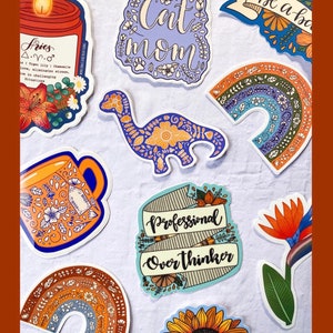 May include: A collection of colorful stickers with various designs, including a candle, a dinosaur, a rainbow, a sunflower, a mug, and a banner that says "Professional Overthinker". The stickers are all decorated with floral patterns and have a vintage aesthetic.
