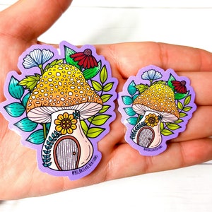 May include: Two purple and white stickers featuring a mushroom house with a yellow and orange mushroom cap, a yellow flower, and green leaves. The text "#MICHAELAELACREATIONS" is on the bottom of the larger sticker.