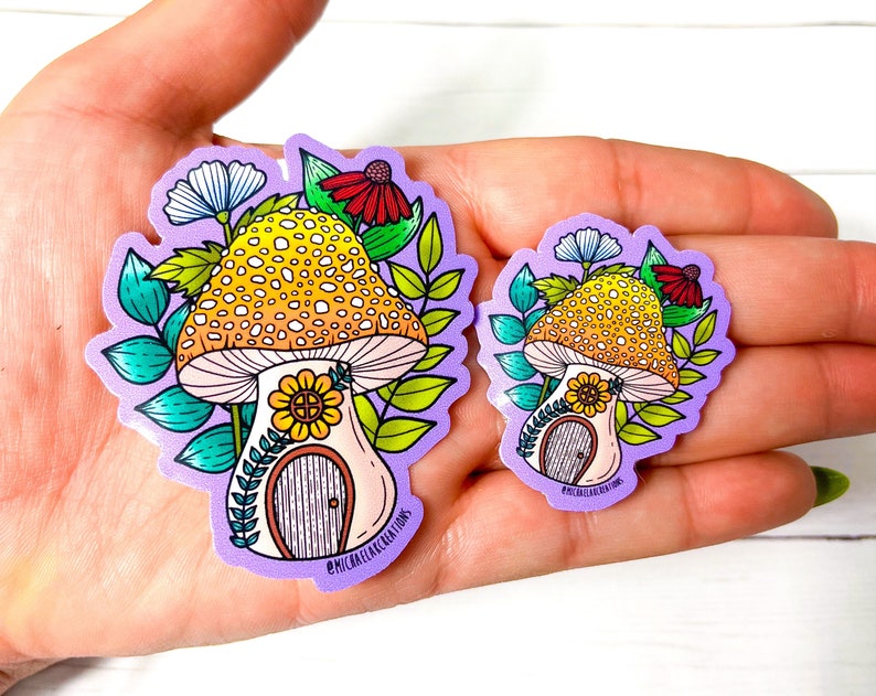 May include: Two purple and white stickers featuring a mushroom house with a yellow and orange mushroom cap, a yellow flower, and green leaves. The text "#MICHAELAELACREATIONS" is on the bottom of the larger sticker.