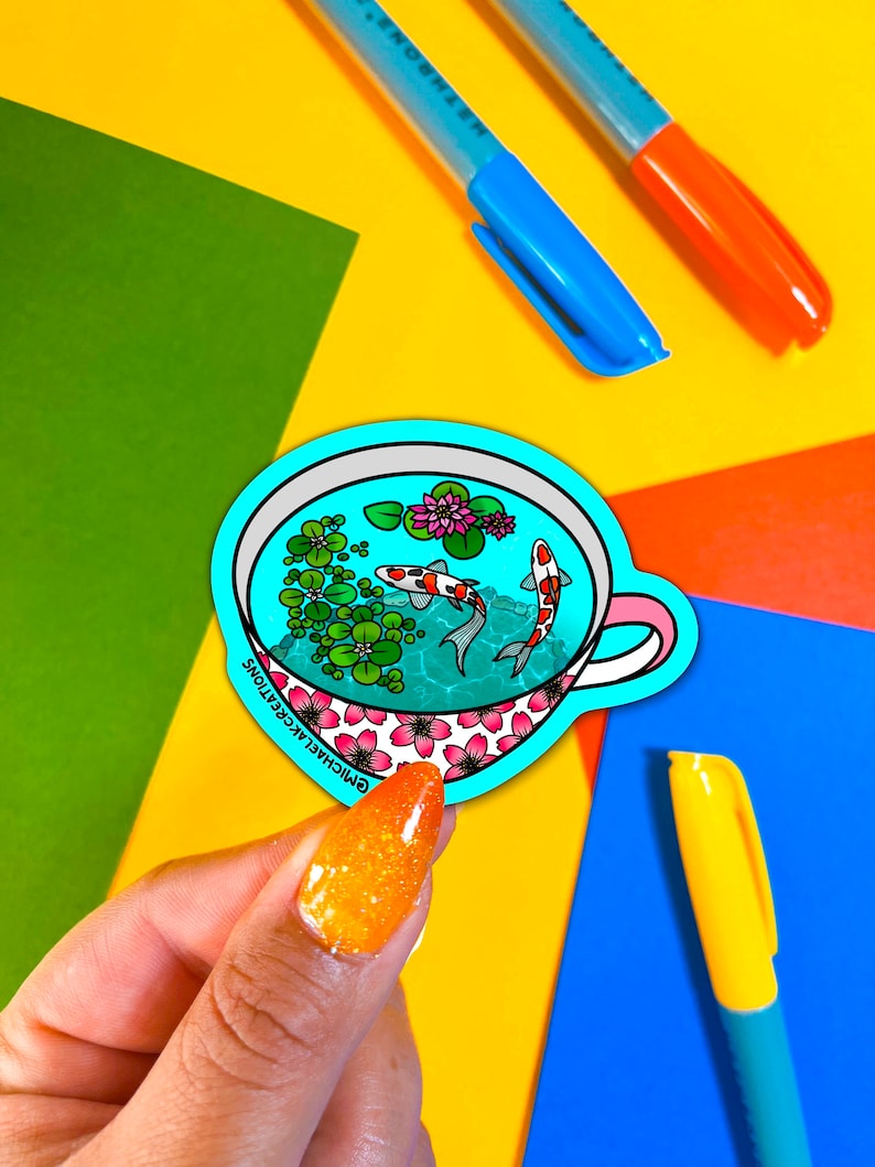 May include: A blue and white sticker with a teacup design. The teacup is filled with water, green plants, and two orange and white koi fish. The text "GMICHALA CREATIONS" is printed on the bottom of the sticker.