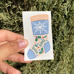 Puede incluir: Una media navide&ntilde;a azul y blanca con un dise&ntilde;o de copo de nieve y hojas de acebo. La media est&aacute; decorada con un borde blanco y un patr&oacute;n rojo y blanco.