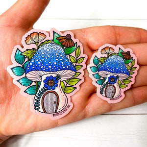 Puede incluir: Dos pegatinas de casa de hongo con sombreros de hongo azules y blancos, hojas verdes y una peque&ntilde;a puerta. La pegatina m&aacute;s grande tiene el texto "@michaelakcreations" en la parte inferior.