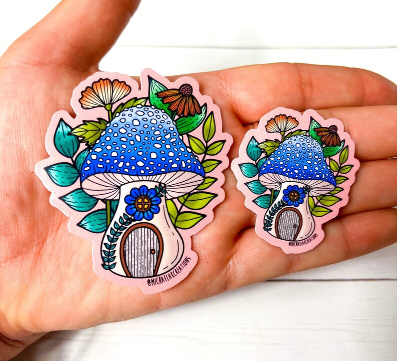 Puede incluir: Dos pegatinas de casa de hongo con sombreros de hongo azules y blancos, hojas verdes y una peque&ntilde;a puerta. La pegatina m&aacute;s grande tiene el texto "@michaelakcreations" en la parte inferior.