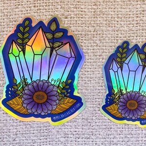 May include: Two iridescent holographic stickers featuring a cluster of blue crystals with a purple flower and green leaves. The larger sticker has the text "@MICHAELACKCREATIONS" at the bottom.