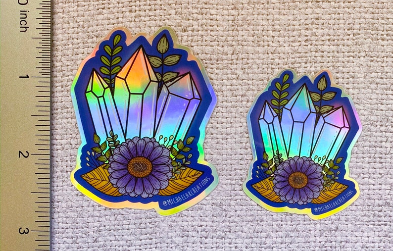 May include: Two iridescent holographic stickers featuring a cluster of blue crystals with a purple flower and green leaves. The larger sticker has the text "@MICHAELACKCREATIONS" at the bottom.