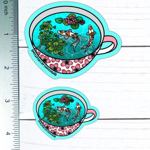 May include: A sticker of a teacup with a blue and white floral pattern. The teacup is filled with water and two orange and white koi fish. The sticker is on a white wooden background.