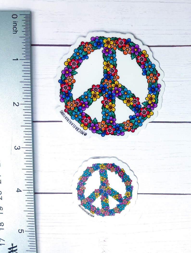 May include: Two peace sign stickers with a floral design. The stickers are white with a peace sign in the center. The peace sign is outlined in black and filled with colorful flowers. The stickers are on a white wooden background.