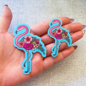 May include: Two pink flamingo stickers with floral designs. The stickers are on a light blue background. The text on the stickers reads "@michaelaellencreations".