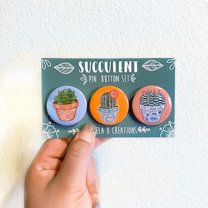 May include: A set of three round succulent pin buttons. The pins are on a teal card with the text "SUCCULENT PIN BUTTON SET". Each button features a different succulent design with text: "SUCC IT", "LOOKIN SHARP", and "YOU GROW GIRL".