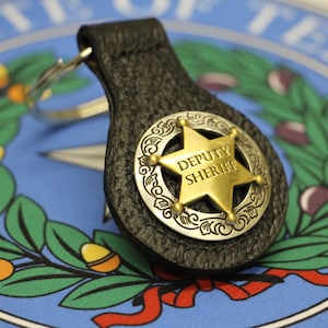 May include: A black leather keychain with a gold and silver sheriff's badge. The badge features a five-pointed star with the words "DEPUTY SHERIFF" inscribed on it.