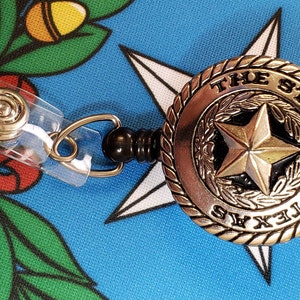 May include: A silver retractable badge holder with a black plastic reel. The badge holder features a round, silver charm with the words "THE STATE OF TEXAS" and a five-pointed star in the center.