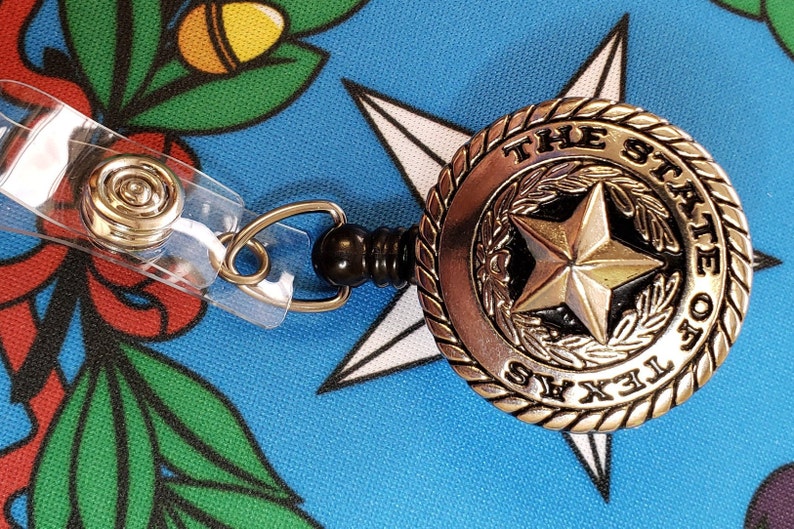 May include: A silver retractable badge holder with a black plastic reel. The badge holder features a round, silver charm with the words "THE STATE OF TEXAS" and a five-pointed star in the center.