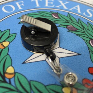 May include: A black retractable badge holder with a silver clip and a clear plastic strap. The badge holder is on top of a blue and white circular design with the words "State of Texas" printed in black.