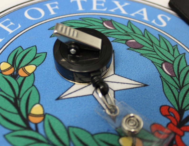 May include: A black retractable badge holder with a silver clip and a clear plastic strap. The badge holder is on top of a blue and white circular design with the words "State of Texas" printed in black.