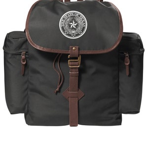 May include: A dark green canvas backpack with brown leather accents. The backpack has a large pocket on the front with a white embroidered seal that reads "The State of Texas".