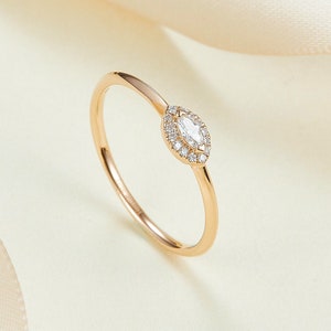 May include: A gold ring featuring a marquise-cut diamond centre stone, encircled by a halo of smaller diamonds. The ring's band is slender and polished, with a warm gold hue. The ring is presented on a cream-coloured surface.