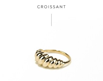 Puede incluir: Un anillo dorado con un diseño único en forma de croissant. El anillo está hecho de metal y tiene un acabado liso y pulido. CROISSANT y PURPLEMAY JEWELLERY están escritos en la imagen.