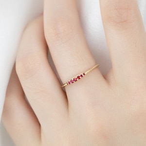 May include: A slender gold ring adorned with a line of tiny, bright red gemstones. The ring's design is minimalist and refined, with the gemstones set closely together. The ring is displayed on a hand, showcasing its elegant simplicity and delicate sparkle.
