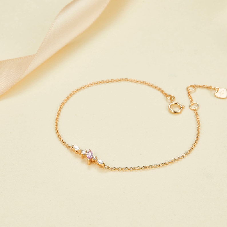 May include: A delicate gold chain bracelet with a pink and white gemstone cluster. The bracelet has a lobster clasp closure.