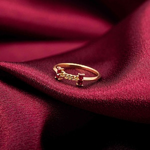 Könnte beinhalten: Ein goldfarbener Ring mit einem kettenartigen Design und zwei rechteckigen roten Edelsteinen. Der Ring ist auf einem tiefroten, seidigen Stoff platziert, was eine luxuriöse und elegante Ästhetik erzeugt.