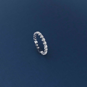 Puede incluir: Un anillo plateado con una fila continua de pequeñas piedras preciosas redondas y transparentes. El anillo se encuentra sobre un fondo azul oscuro, creando un contraste que resalta el brillo de las piedras. Es una joya delicada.