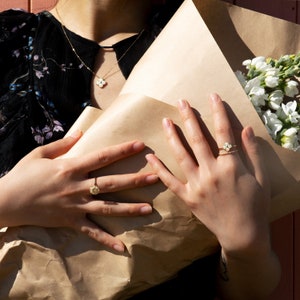 May include: A woman's hands holding a bouquet of white flowers wrapped in brown paper. She is wearing a gold necklace with a flower pendant and two gold rings on her fingers.